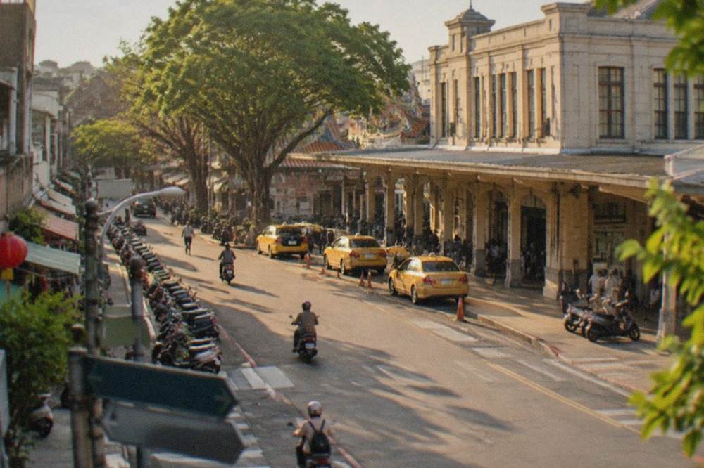 A Tainan street corner on a sunny day, low shophouses and scooters, direction boards visible but turned away and out of focus