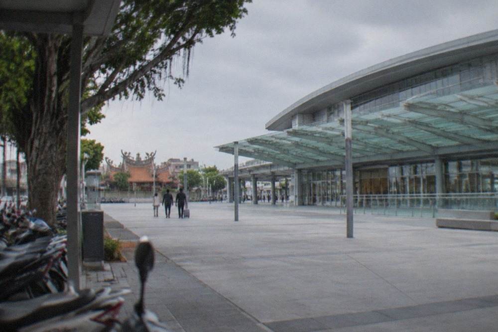 A modern high speed rail station exterior with a glass canopy and a wide open plaza in front