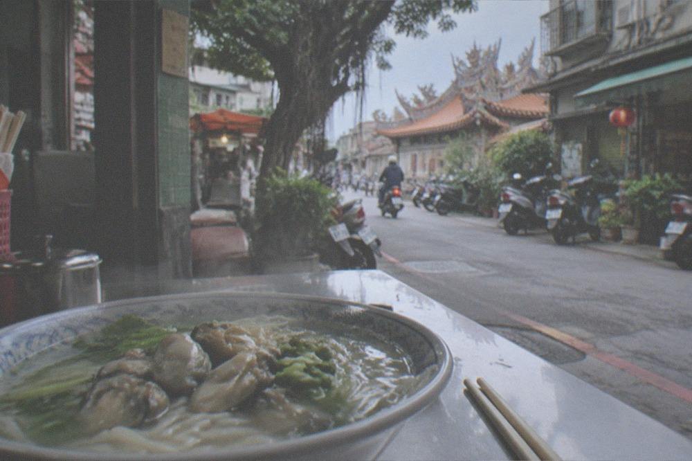 A steaming bowl of noodle soup with fresh oysters on a small restaurant table beside chopsticks