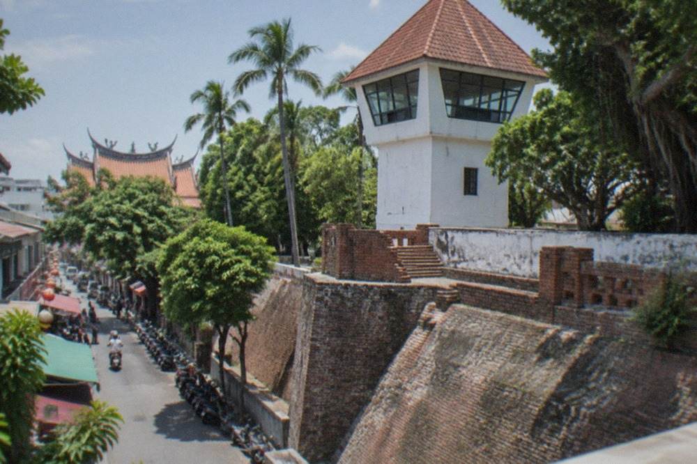 A white-walled fort lookout tower with a pointed red-tiled roof above old brick ramparts and palm trees