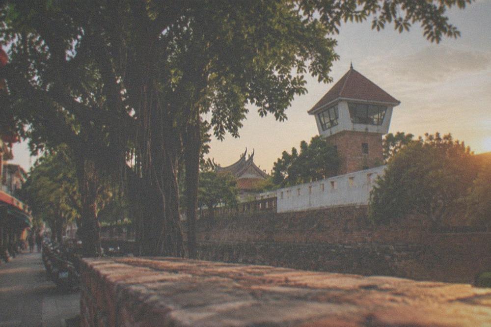 A lookout tower with a pointed red roof seen over a low brick wall and trees