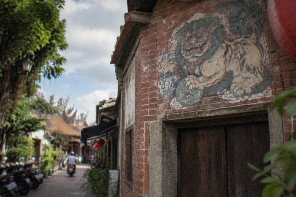 A weathered sword lion painting on a red brick wall above a wooden door in a narrow Anping lane