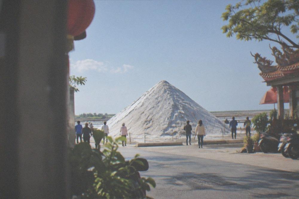 A white salt mountain rising above flat coastal terrain with visitors walking near its base
