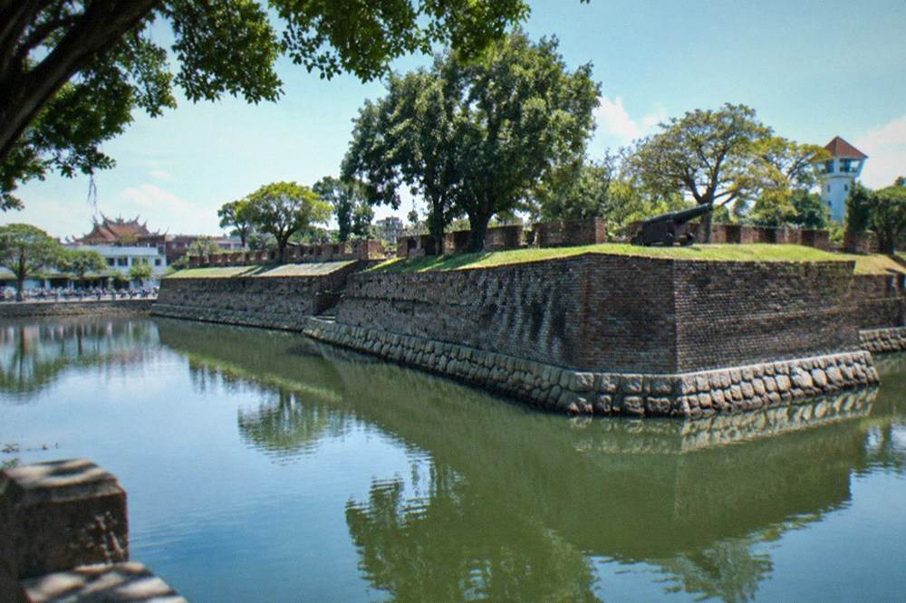 A wide water moat reflecting trees, encircling grassy fort ramparts on a clear afternoon