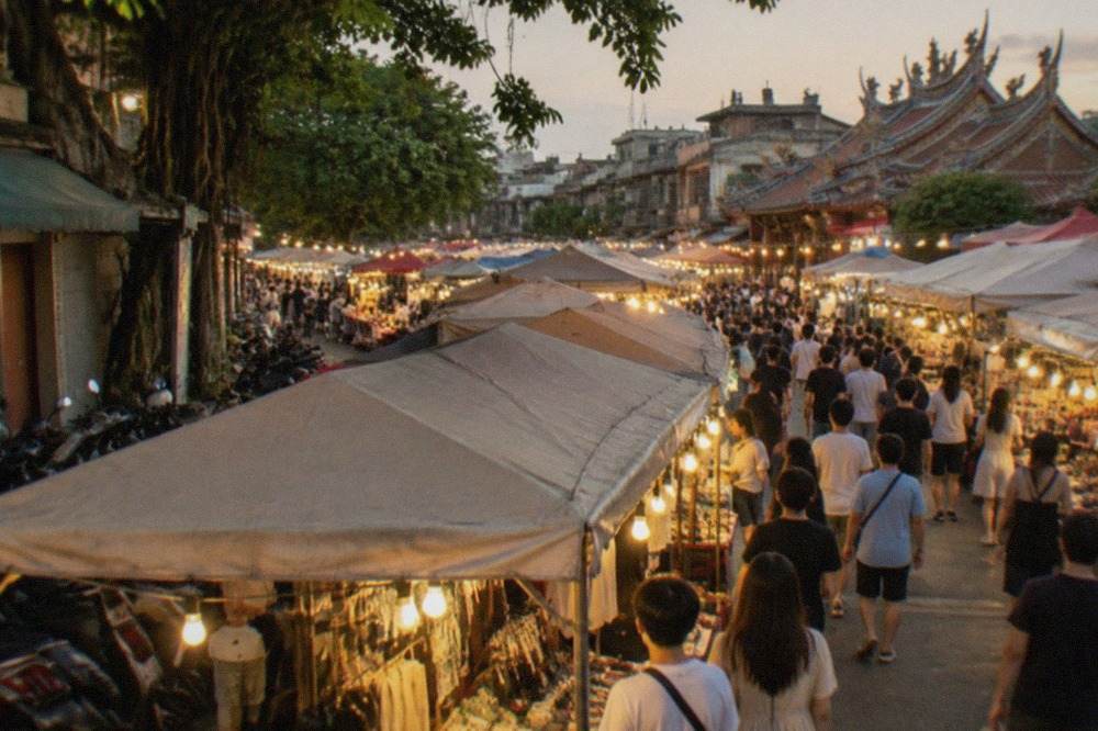 A crowded open-air night market aisle lit by rows of yellow stall lights at dusk