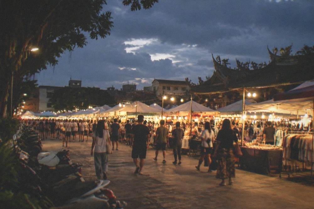 A wide view of a night market ground with lit stalls and crowds under a dark sky