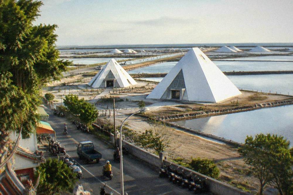 A flat coastal road running between fish ponds toward distant salt piles under open sky