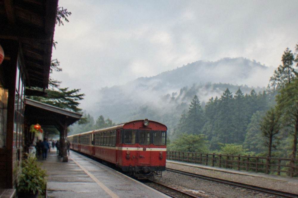 A mountain railway platform with a narrow-gauge train waiting as mist rolls over forested ridges