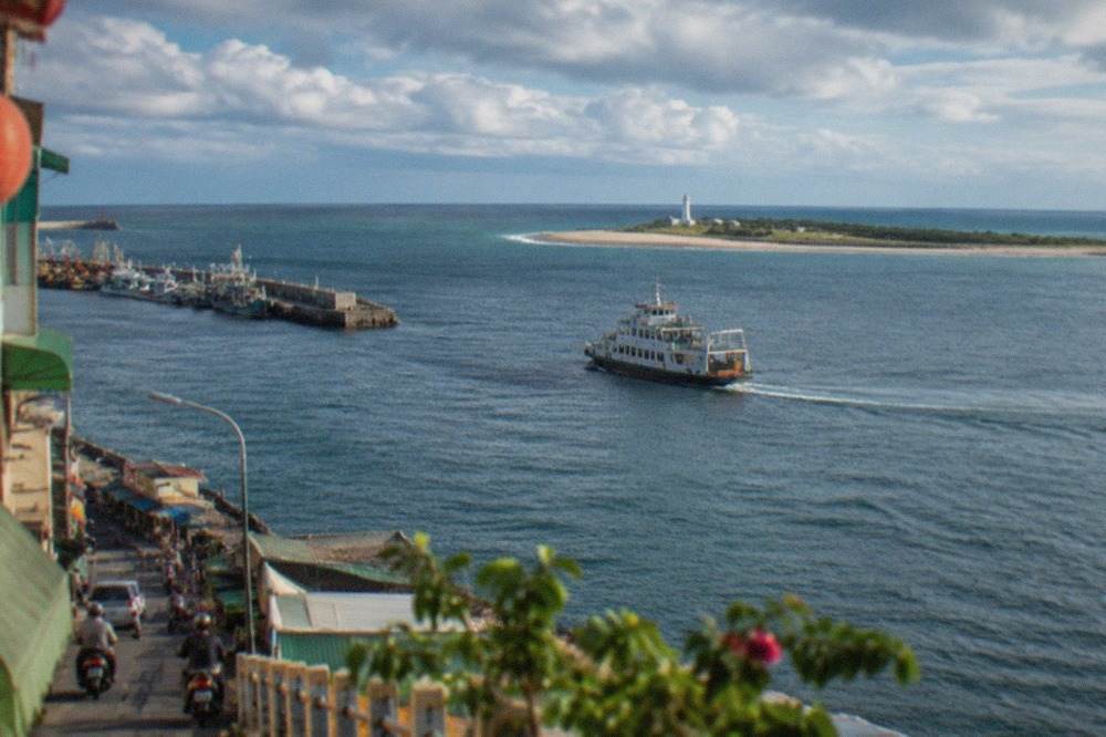 A ferry crossing a wide harbor toward a long narrow island with a beach and a white lighthouse in the distance