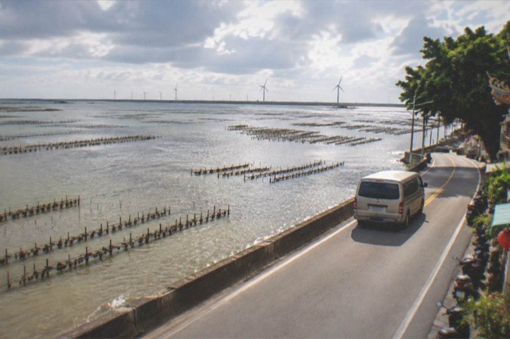 A tour van driving along a coastal road beside lagoon water and wind turbines