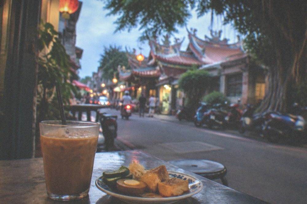 A glass of iced coffee on a table beside a plate of Taiwanese snacks