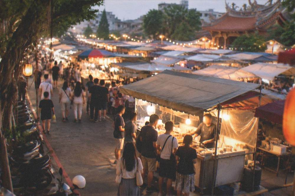 A single food stall glowing under a bright bulb at night, with a short queue of customers waiting at the counter