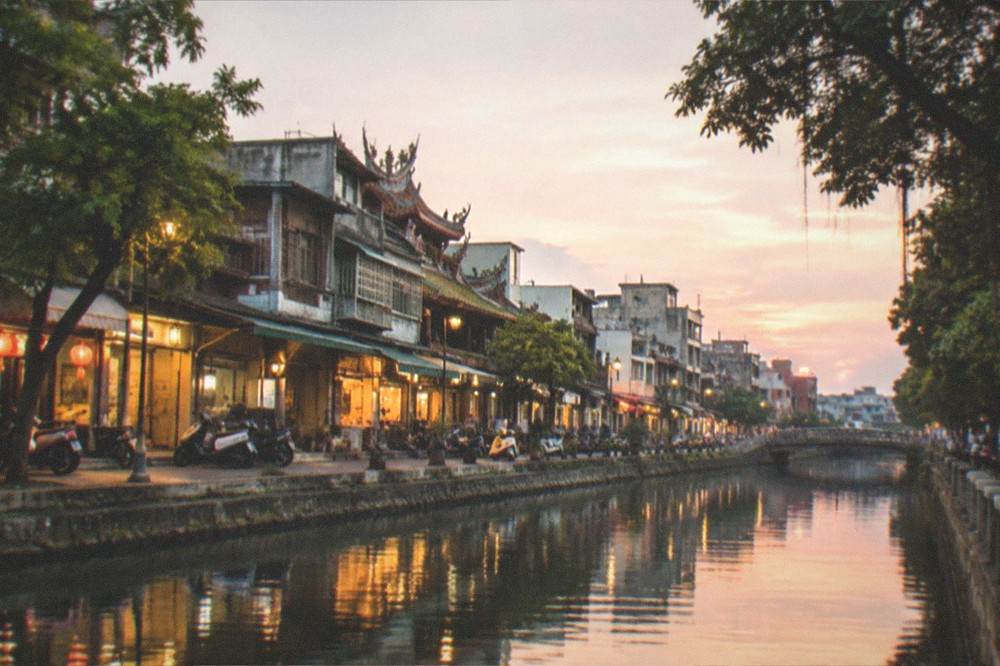 A canal-side street in Tainan at dusk with lit shopfronts reflected in the water