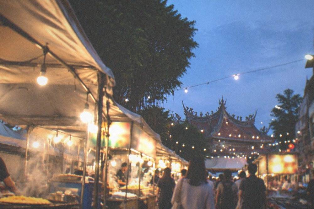 Crowded stalls at a Tainan night market in the evening, bare bulbs overhead and steam above the griddles, the lit boards above the stalls out of focus